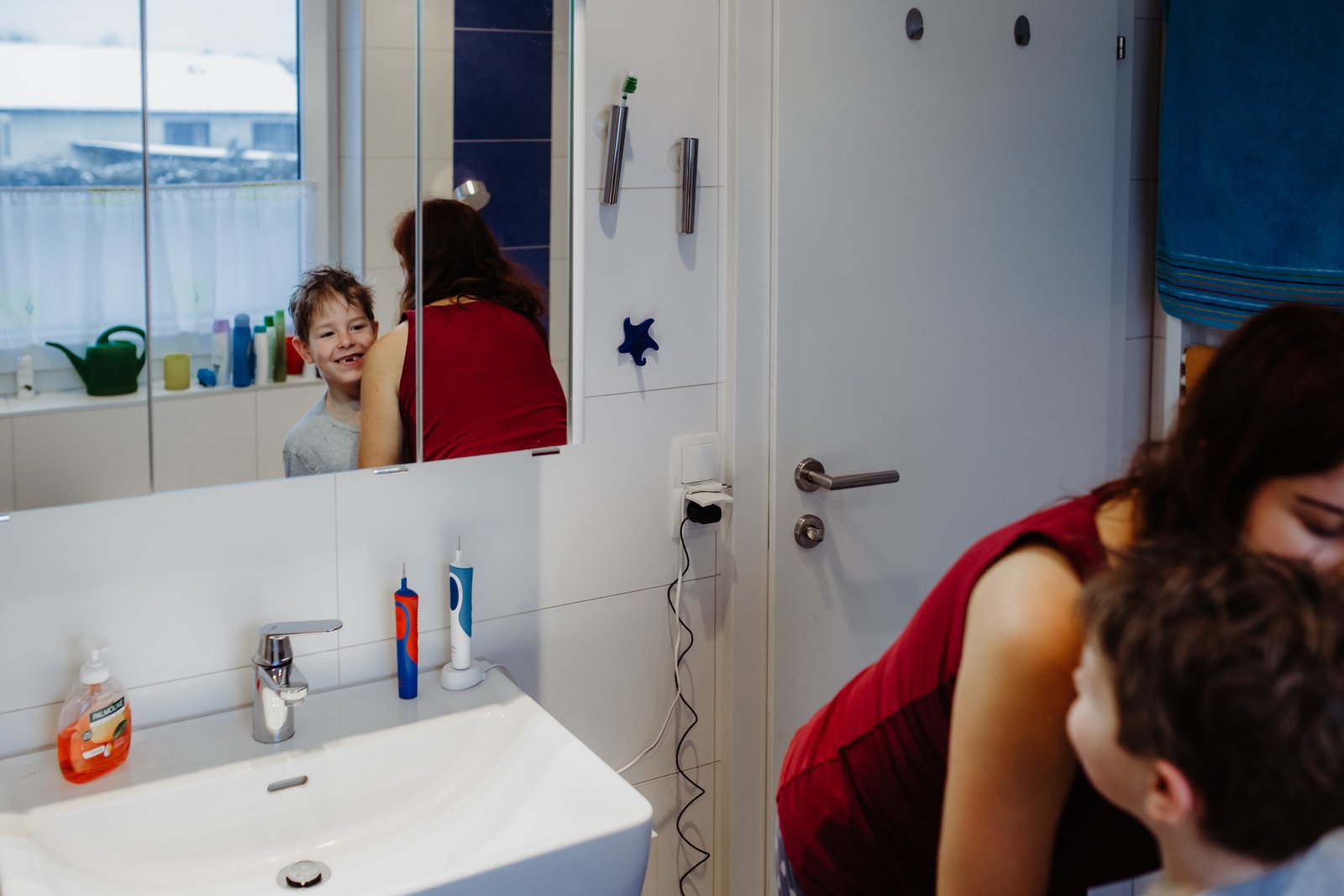 Ein Junge grinst im Badezimmerspiegel, während seine Mutter neben ihm steht.